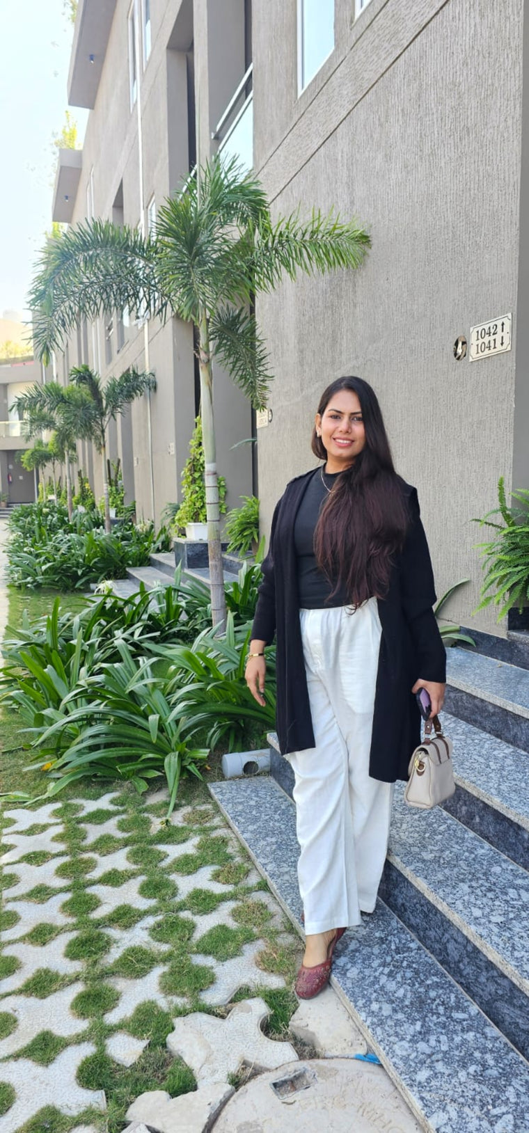 A woman stands on outdoor stone steps in front of a modern building, wearing a black blazer over a black top and white wide-leg pants, holding a small beige handbag.